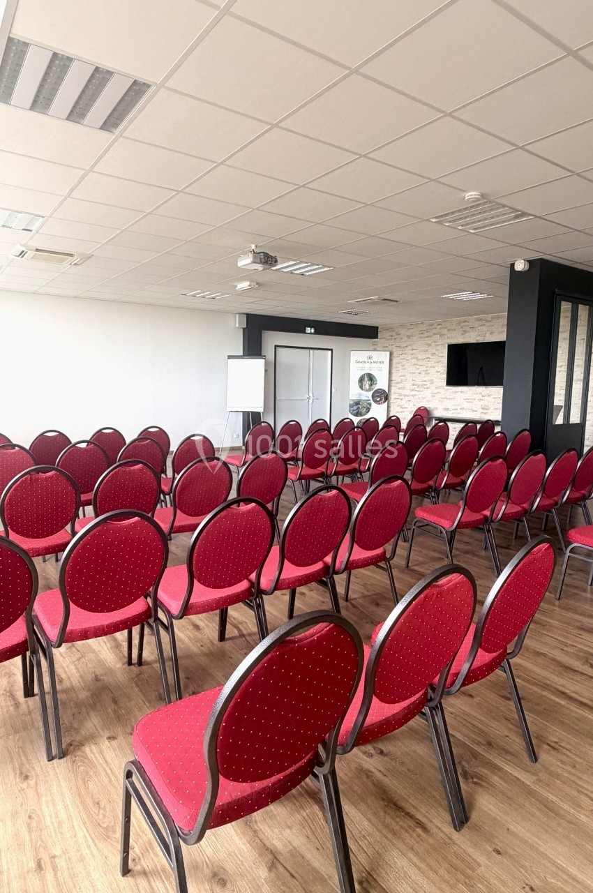 Salle de conférence avec des rangées de chaises rouges alignées sur un sol en bois, écran et tableau blanc en arrière-plan.