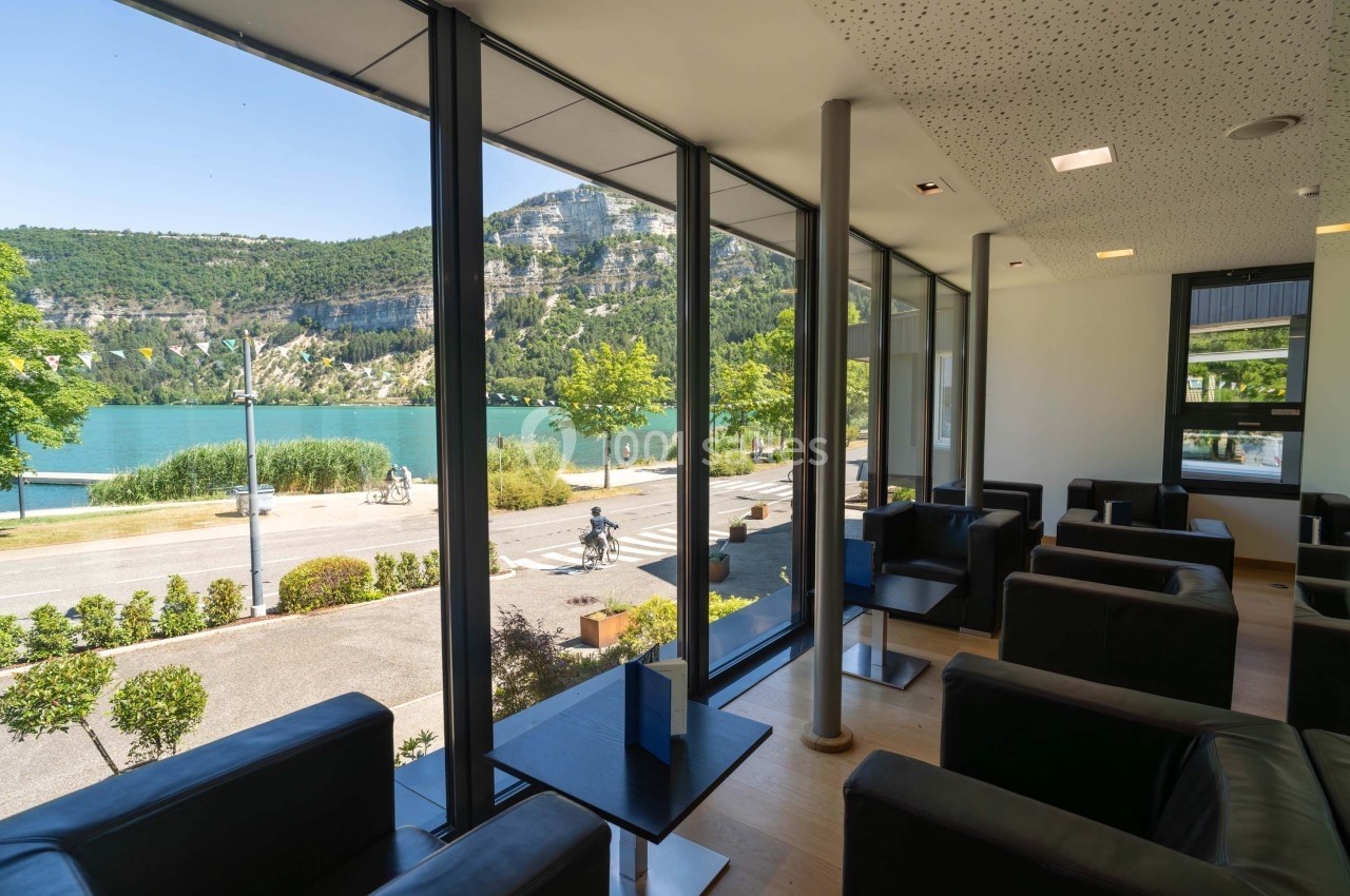 Salle lumineuse avec fauteuils noirs, grandes baies vitrées offrant une vue sur un lac, des arbres et des collines…