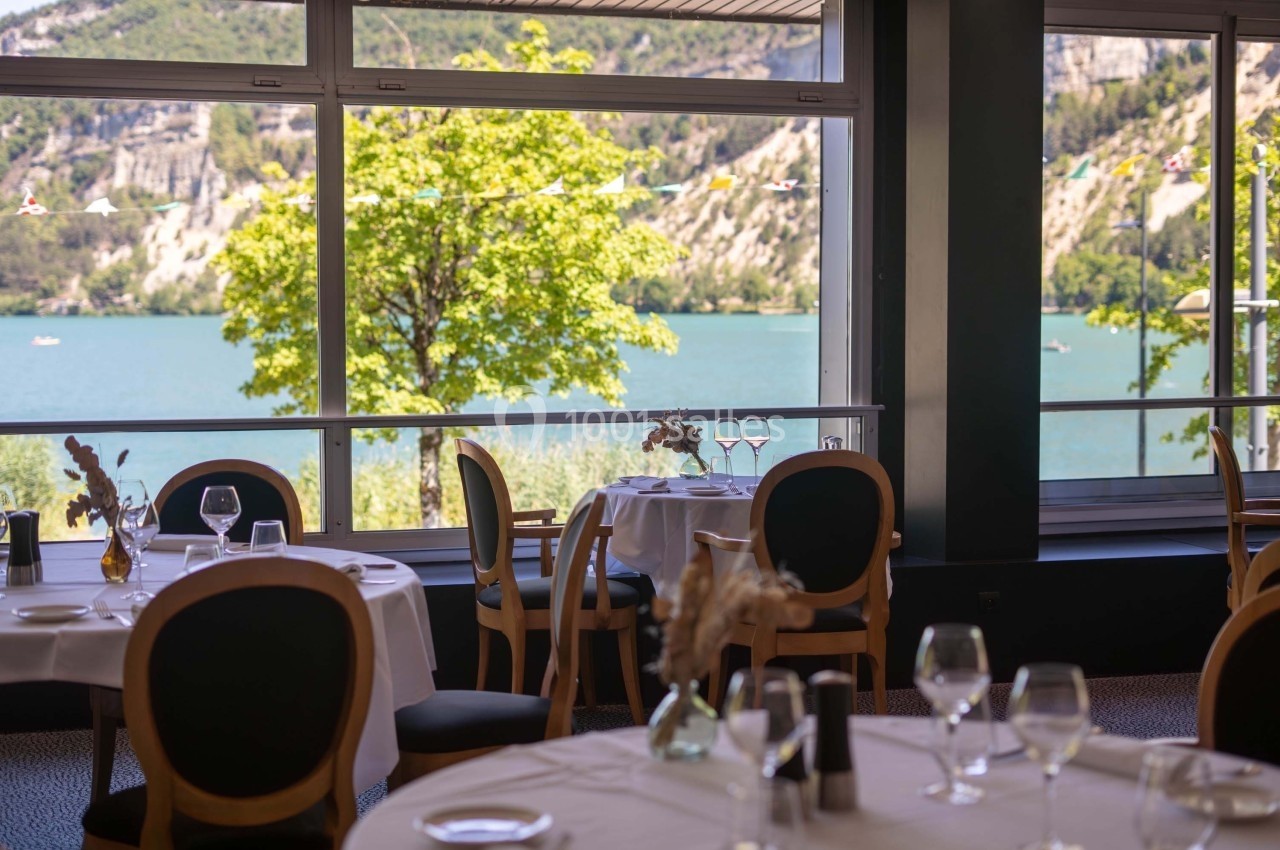 Salle de restaurant lumineuse avec vue sur un lac, des montagnes et des tables dressées près de grandes baies vitrées.