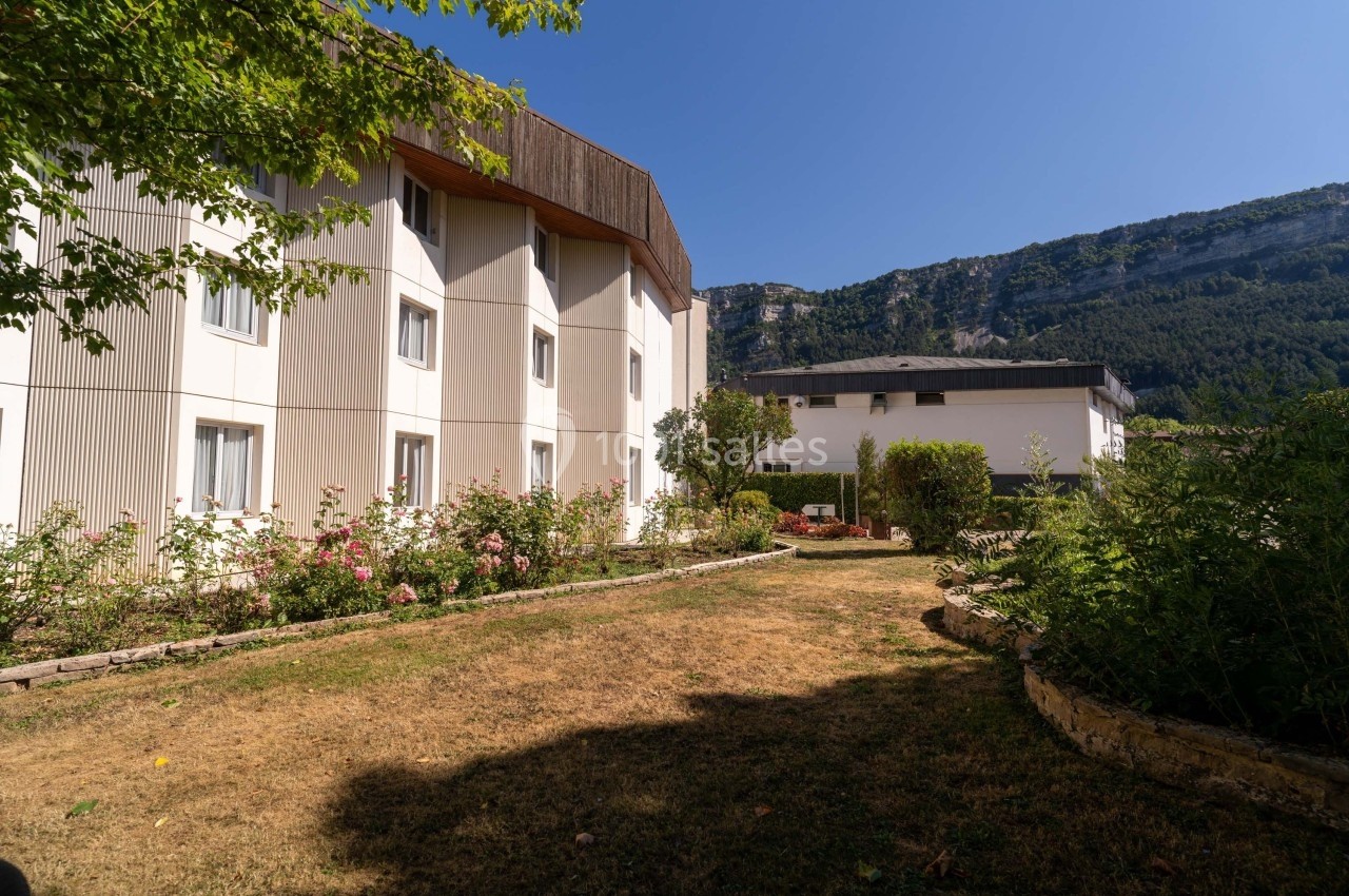 Bâtiment moderne entouré d'un jardin avec des arbustes et des fleurs, situé au pied d'une montagne sous un ciel bleu.