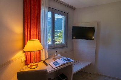 Chambre d'hôtel avec lit double, bureau, lampe, télévision murale et fenêtre donnant sur un paysage verdoyant.