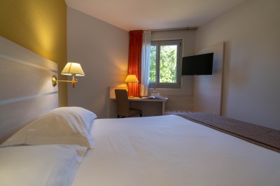 Chambre d'hôtel avec lit double, bureau, lampe, télévision murale et fenêtre donnant sur un paysage verdoyant.