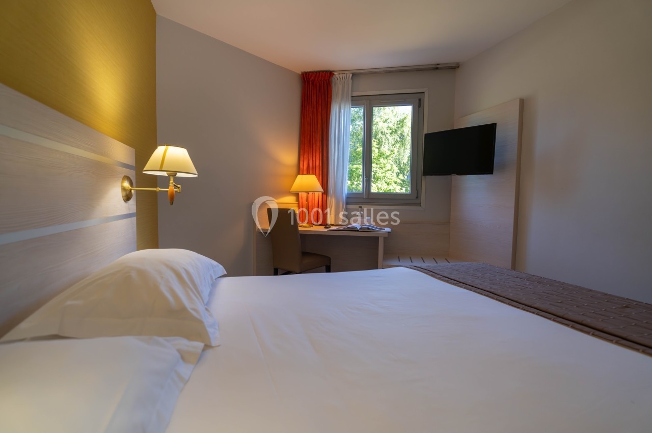 Chambre d'hôtel avec lit double, bureau, lampe, télévision murale et fenêtre donnant sur un paysage verdoyant.