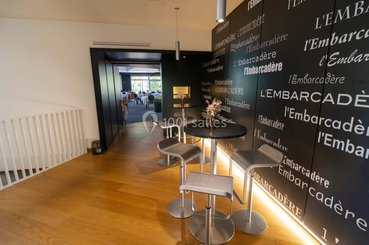 Espace intérieur moderne avec table haute, tabourets et mur noir décoré de mots répétés, éclairage chaleureux.