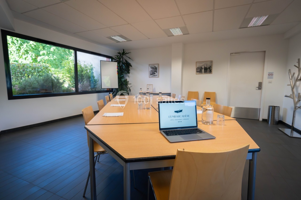 Salle de réunion lumineuse avec une grande table en bois, des chaises, un ordinateur portable et des documents disposés.