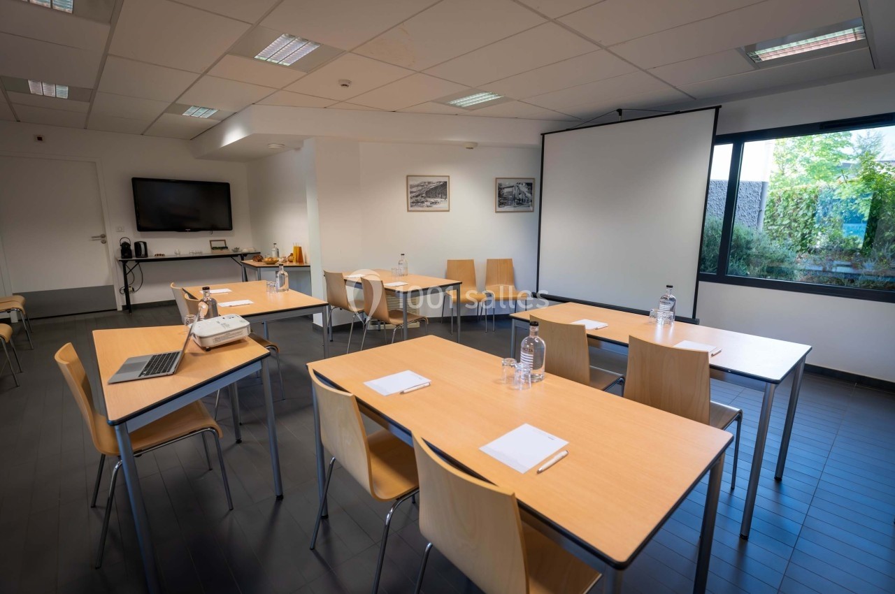 Salle de réunion lumineuse avec tables en bois, chaises, projecteur, écran blanc et vue sur un jardin.