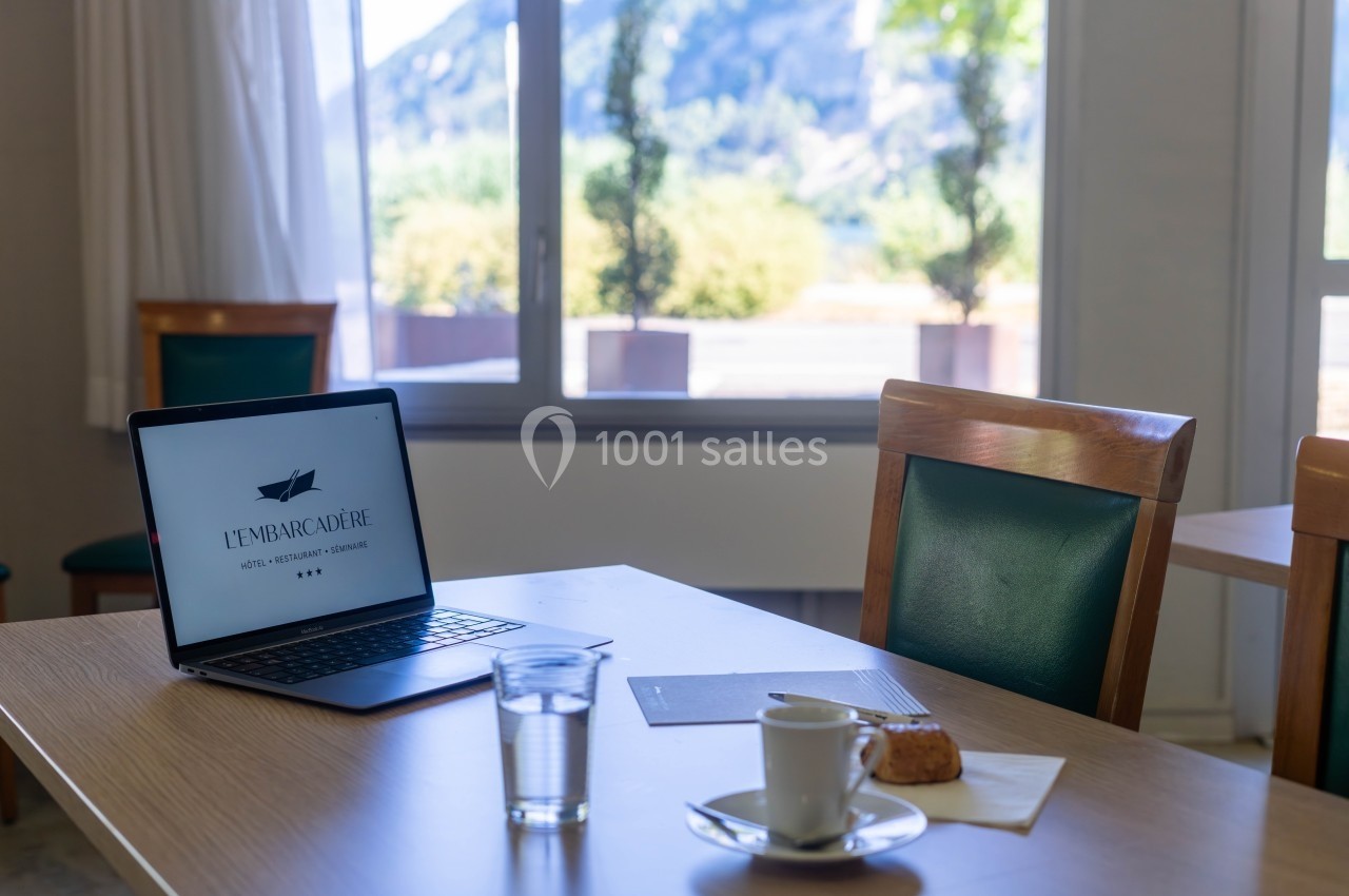 Ordinateur portable, tasse de café, verre d'eau et viennoiserie sur une table en bois près d'une fenêtre lumineuse.