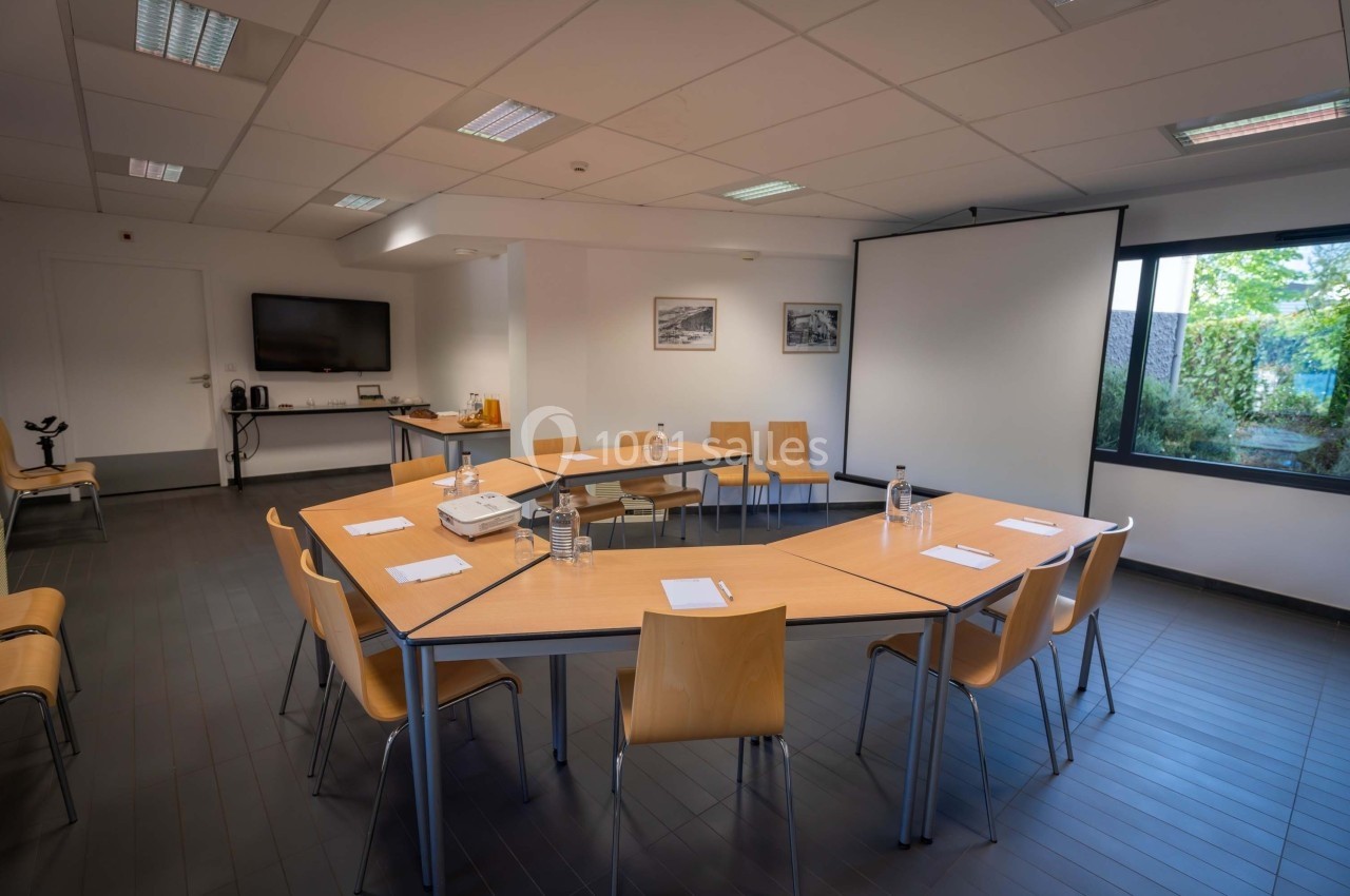 Salle de réunion lumineuse avec tables disposées en U, chaises en bois, écran de projection et matériel de présentation.