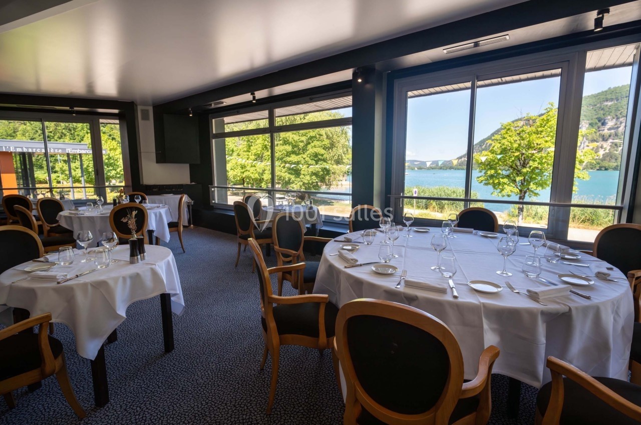 Salle de restaurant lumineuse avec tables dressées, grandes baies vitrées offrant une vue sur un lac et des collines…