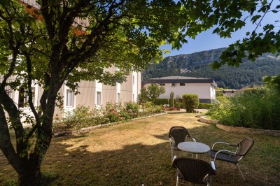 Chambre lumineuse avec lit double, bureau, chaise, télévision murale et fenêtre donnant sur un espace vert.