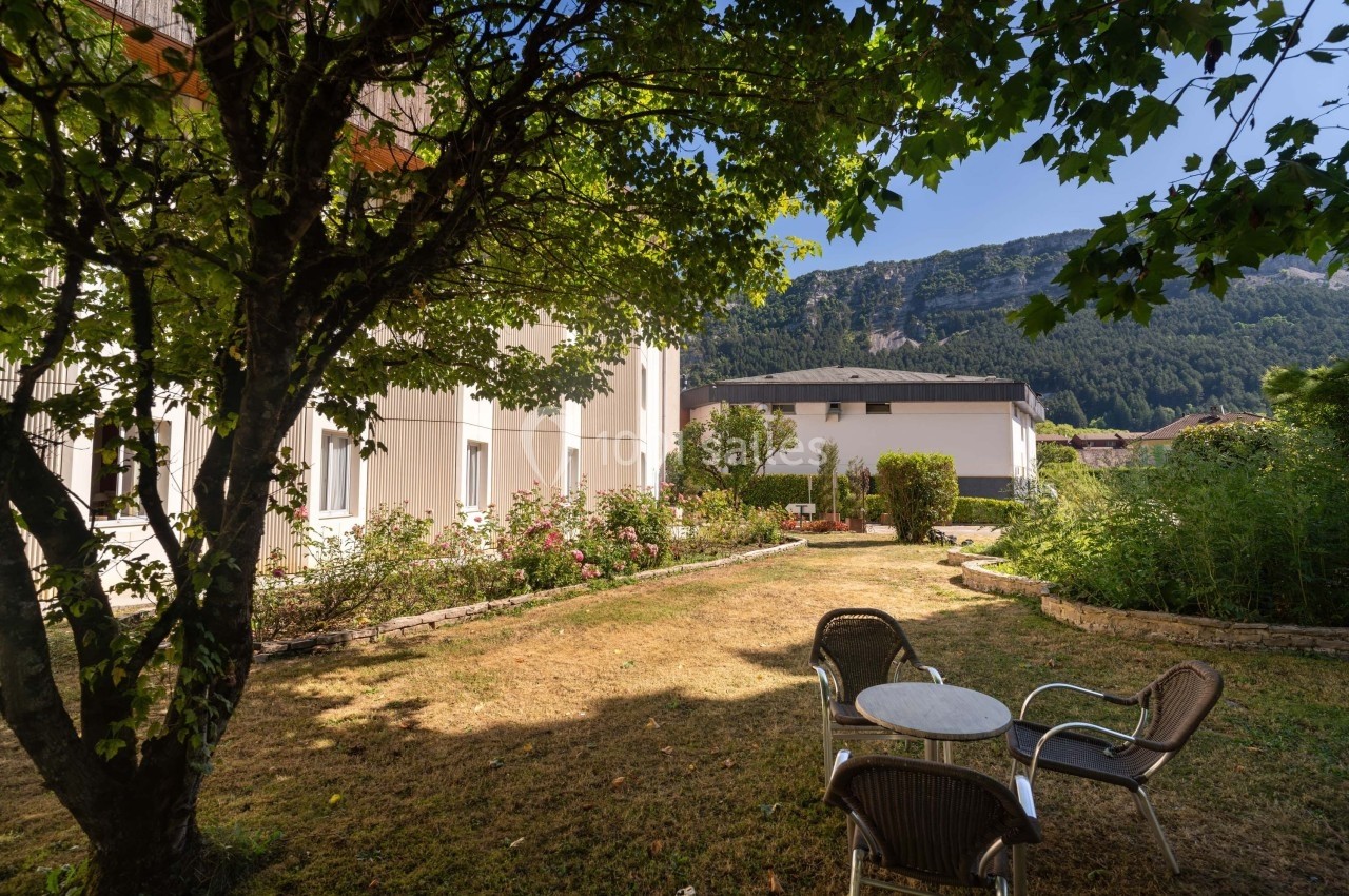 Jardin avec pelouse, arbres, table et chaises, entouré de bâtiments et montagnes en arrière-plan.