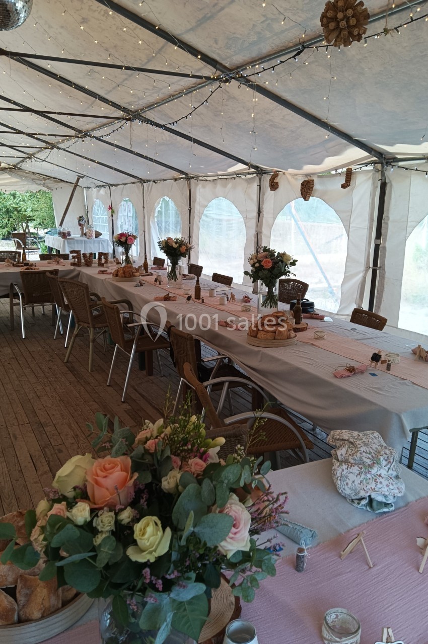 Salle de réception sous chapiteau avec tables décorées de nappes, fleurs et lumières suspendues.