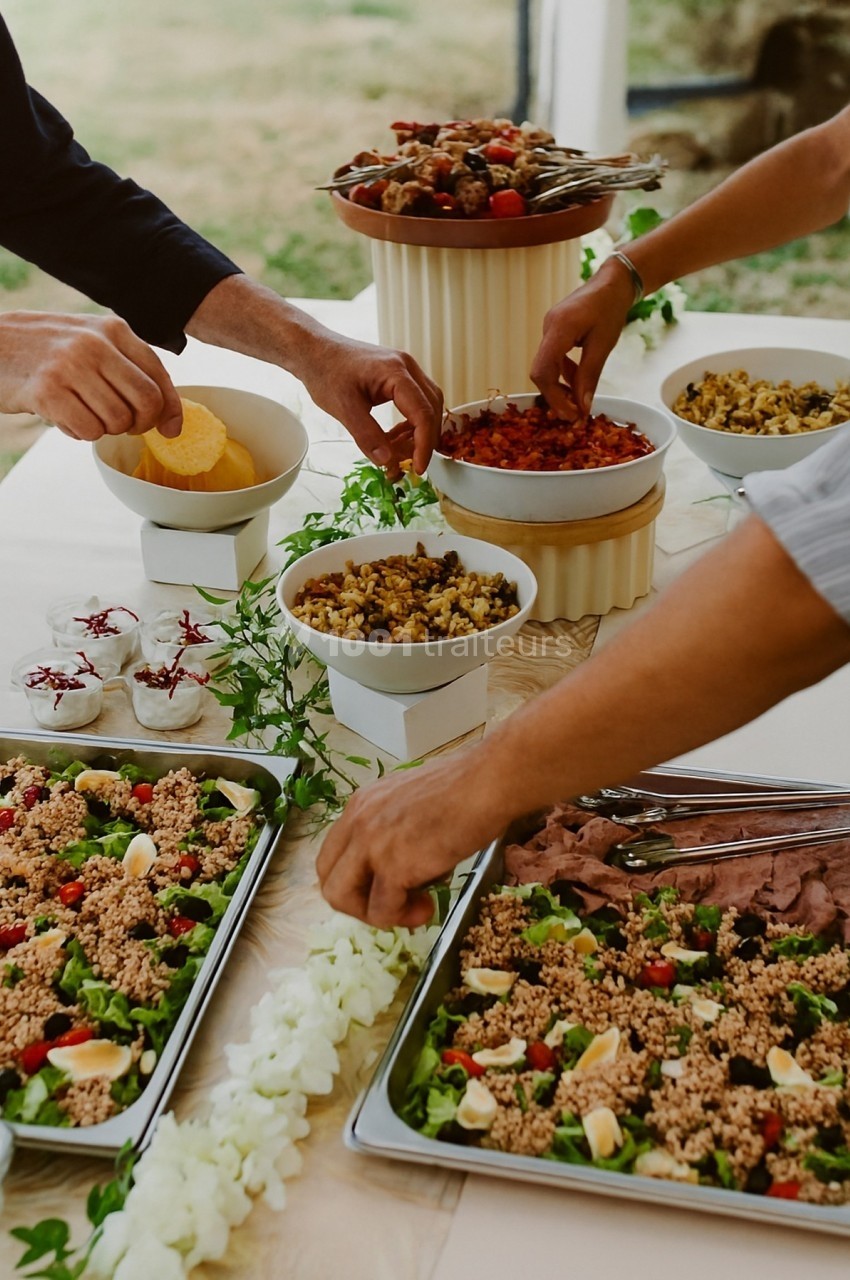 Des mains se servent de plats variés sur une table garnie de salades, légumes, viandes et desserts.