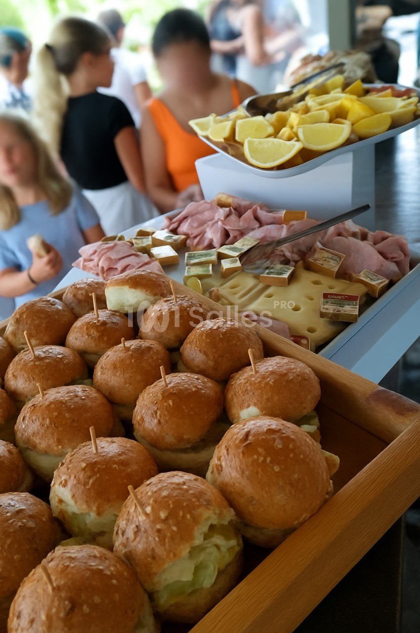 Plateau de petits pains garnis, charcuterie, fromage et quartiers de citron, avec des personnes en arrière-plan flou.