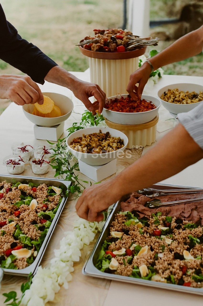 Des mains servent des plats variés sur une table garnie de salades, légumes, viandes et desserts.