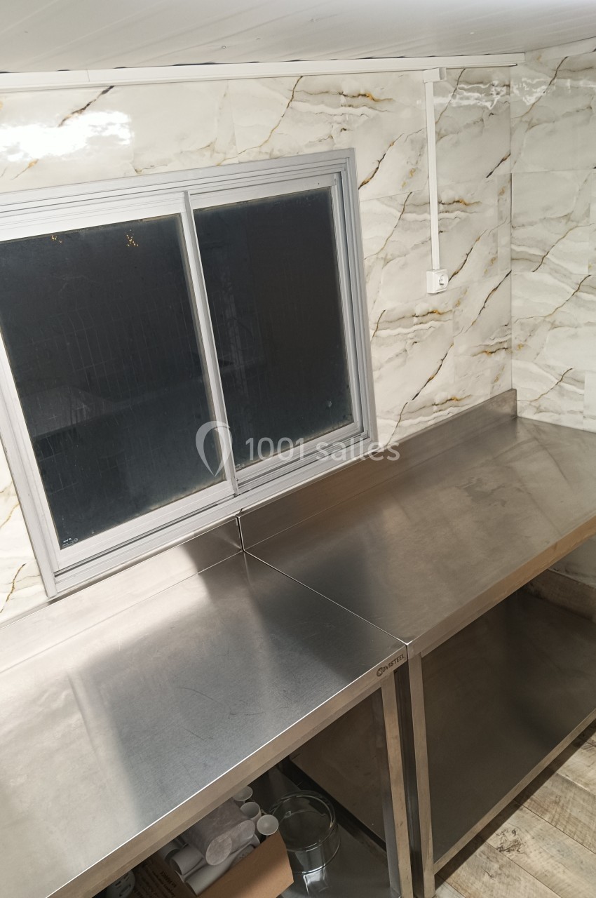 Plan de travail en acier inoxydable sous une fenêtre à double vitrage, dans une pièce aux murs carrelés effet marbre.