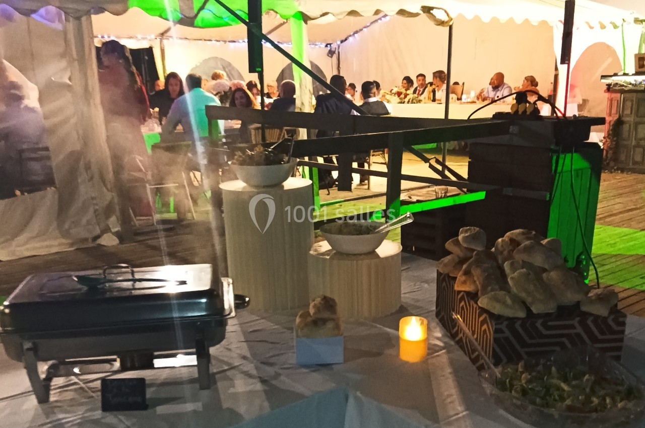 Buffet en extérieur sous une tente éclairée, avec des plats, du pain et des convives assis en arrière-plan.