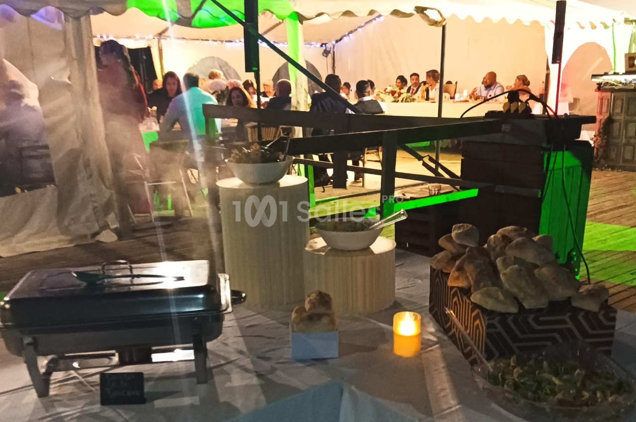 Buffet en extérieur sous une tente éclairée, avec des plats, du pain et des convives assis en arrière-plan.