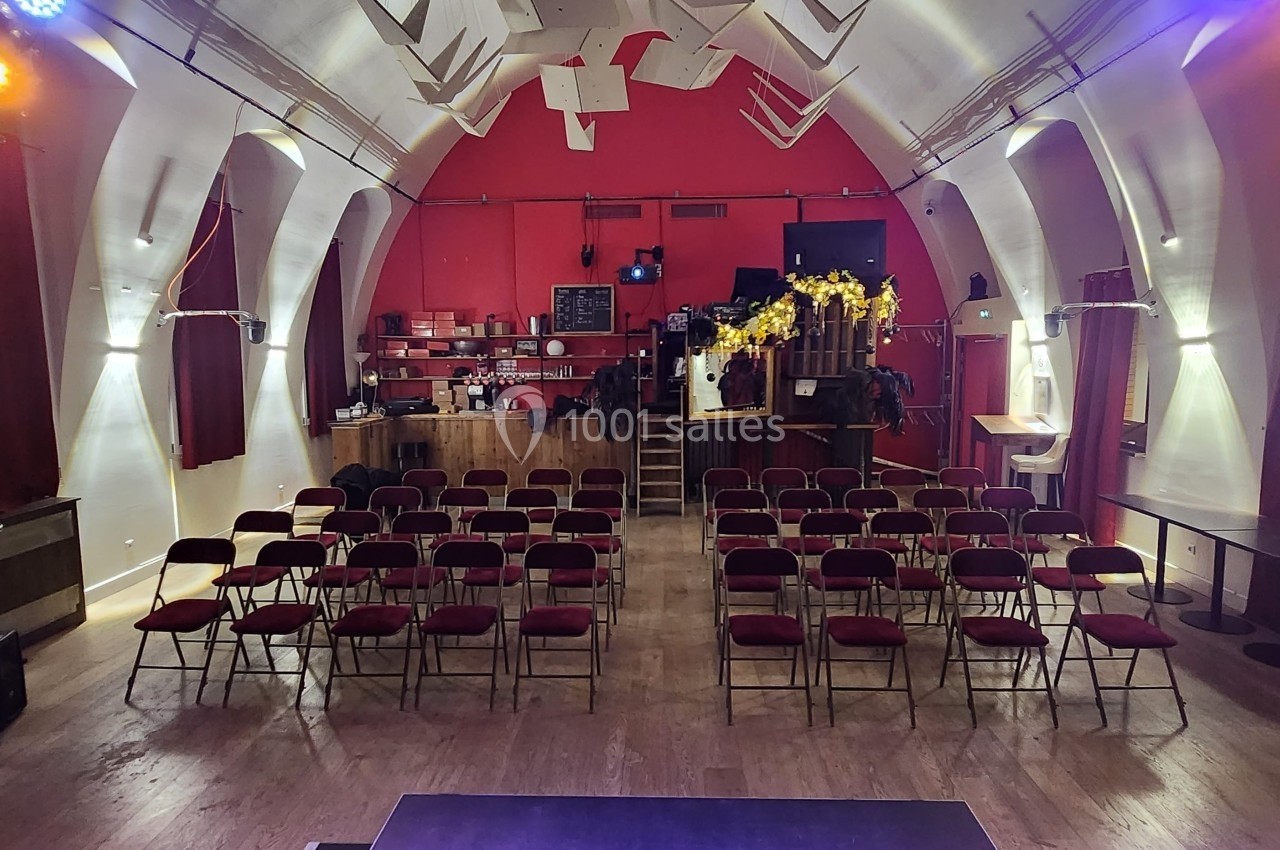 Salle de spectacle avec rangées de chaises rouges, scène au premier plan et bar en fond dans un décor rouge et blanc.