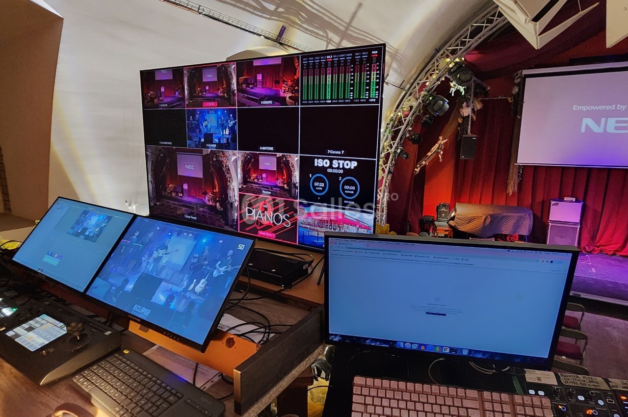 Poste de régie audiovisuelle avec écrans de contrôle, matériel technique et vue sur une scène de théâtre éclairée.
