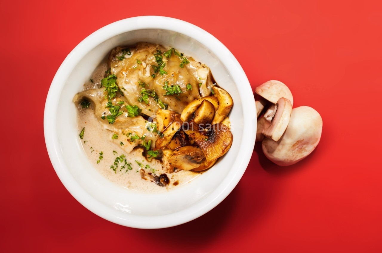 Raviolis dans une sauce crémeuse, garnis de champignons poêlés et de persil, sur fond rouge.