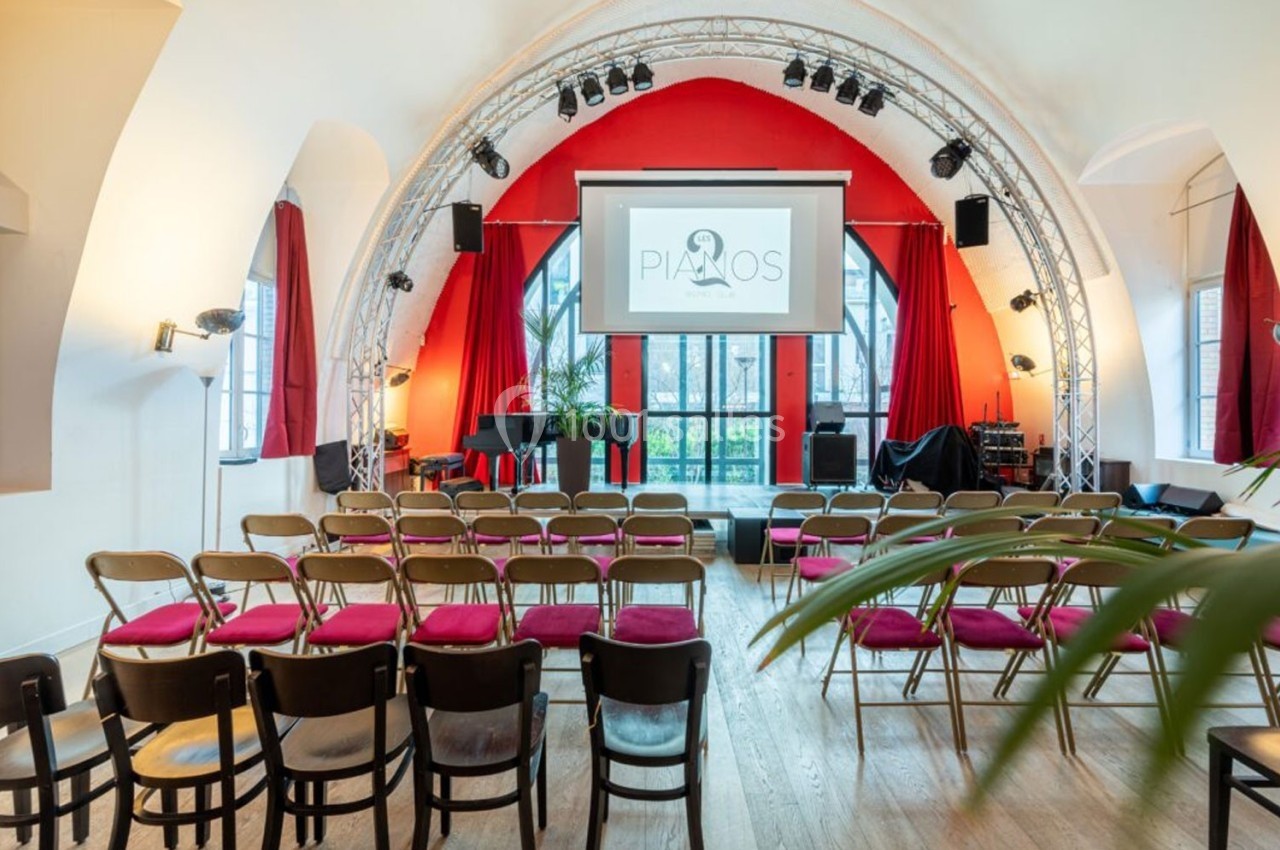 Salle de spectacle avec chaises alignées, scène équipée d'un piano, projecteur et rideaux rouges encadrant une arche.