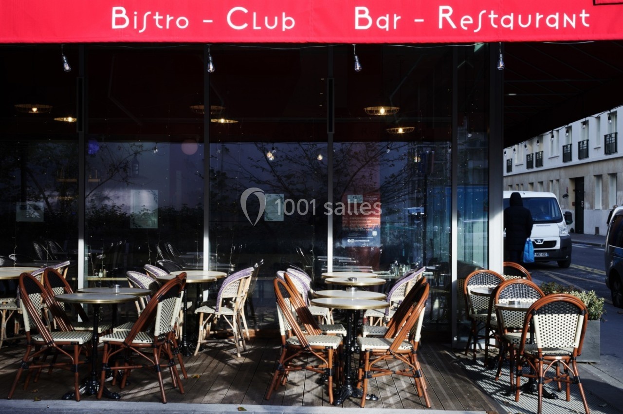 Terrasse extérieure d'un bistro avec des tables et chaises en osier sous un auvent rouge, vue sur la rue adjacente.