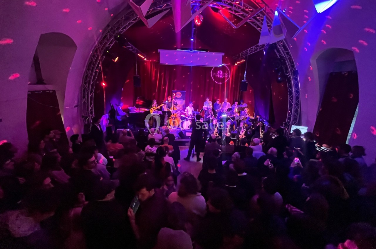 Salle de concert bondée avec un groupe de musiciens sur scène, éclairée par des lumières colorées et une ambiance festive.