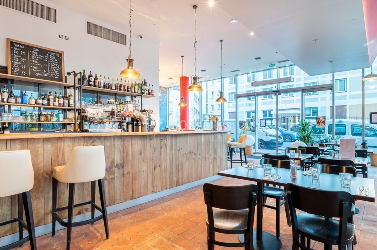 Intérieur d'un café moderne avec un bar en bois, des tables et chaises noires, éclairé par des suspensions lumineuses.