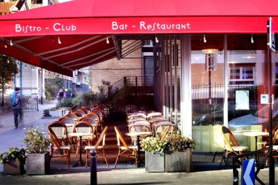 Façade d'un café-restaurant avec terrasse extérieure, tables et chaises sous un auvent rouge marqué ’Les 2 Pianos’.