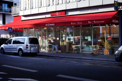 Façade d'un café-restaurant avec terrasse extérieure, tables et chaises sous un auvent rouge marqué ’Les 2 Pianos’.