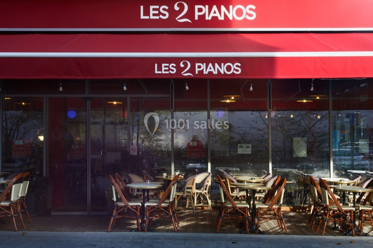 Façade d'un café-restaurant avec terrasse extérieure, tables et chaises sous un auvent rouge marqué ’Les 2 Pianos’.