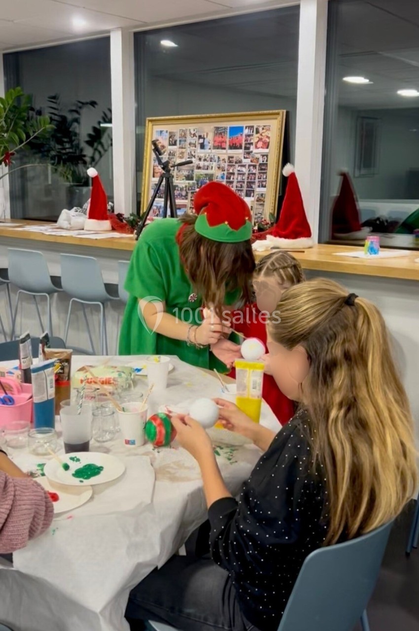 Des enfants et un adulte décorent des boules de Noël dans une salle lumineuse avec des décorations festives.