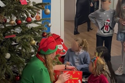 Un Père Noël assis tenant un cadeau, accompagné d'une assistante en tenue d'elfe près d'un sapin décoré.