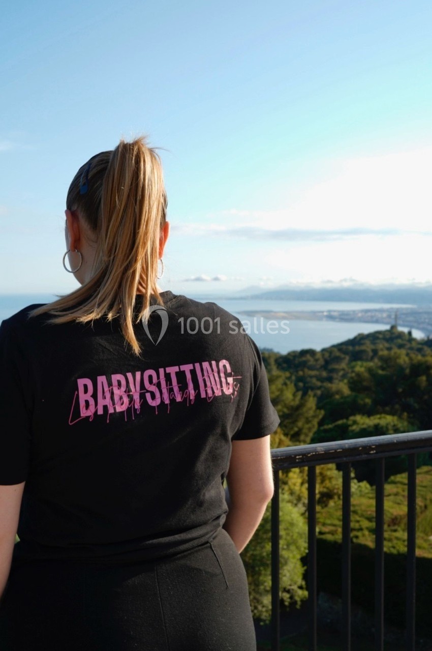 Une femme de dos, portant un t-shirt noir avec l'inscription ’Babysitting’, devant un paysage côtier.