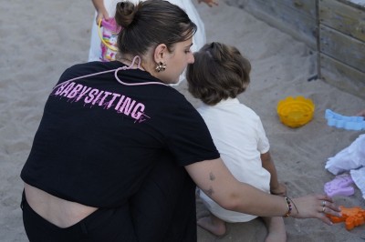 Une femme surveille deux jeunes enfants jouant avec des jouets dans un bac à sable en plein air.