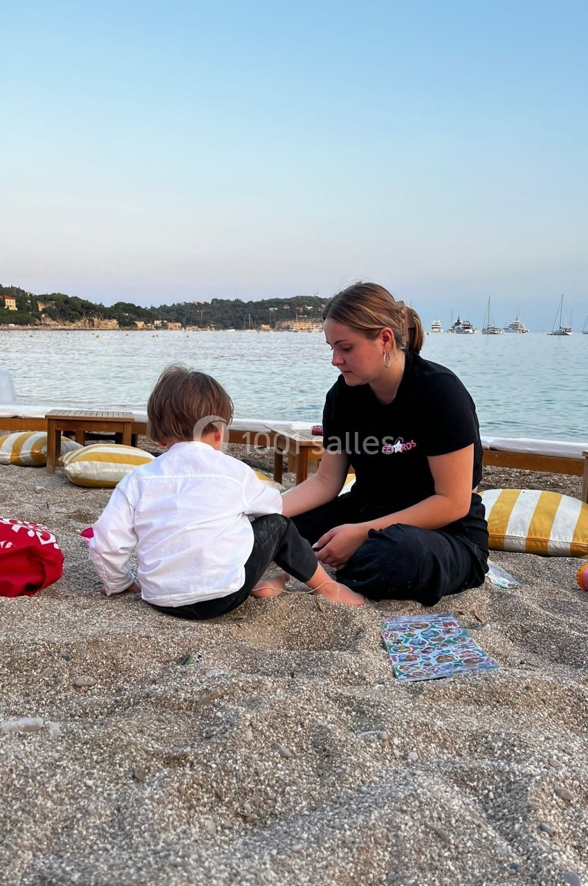 Une femme assise sur une plage joue avec un jeune enfant, avec des bateaux visibles au loin sur l'eau calme.