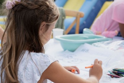 Panneau ’Kids Area’ en bois près d'une aire de jeux, avec une femme de dos portant un t-shirt ’Babysitting’.