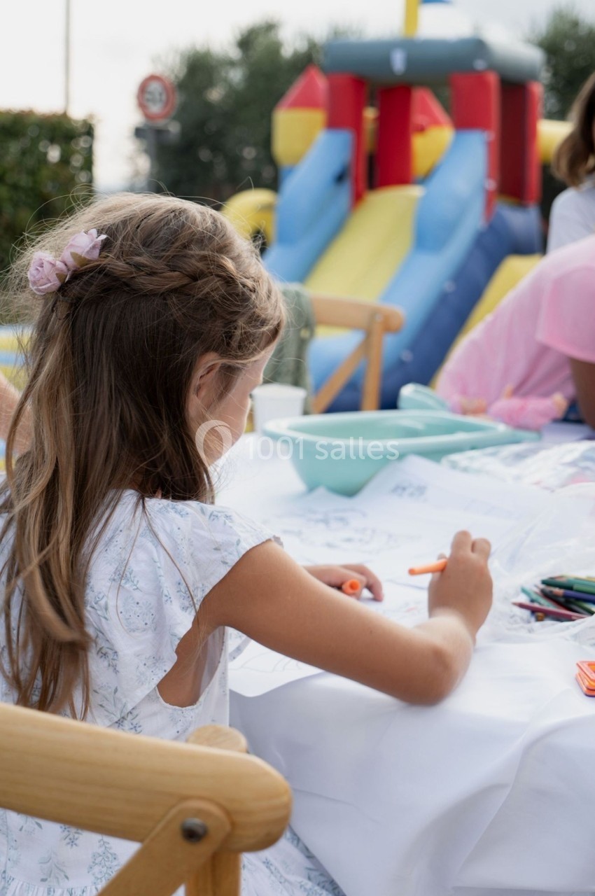 Une fillette dessine avec des crayons de couleur à une table en extérieur, près d'un château gonflable.