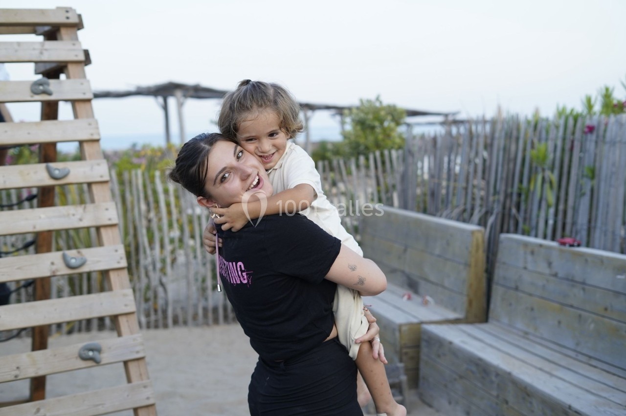 Une femme souriante porte un enfant dans ses bras sur une plage avec des structures en bois en arrière-plan.
