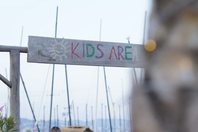 Panneau ’Kids Area’ en bois près d'une aire de jeux, avec une femme de dos portant un t-shirt ’Babysitting’.