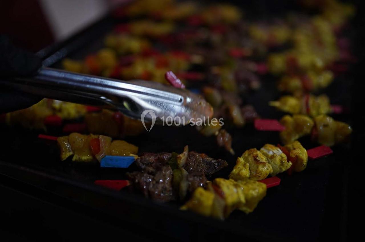 Brochettes de viande et légumes colorés grillées sur une plaque chauffante, manipulées avec une pince en métal.