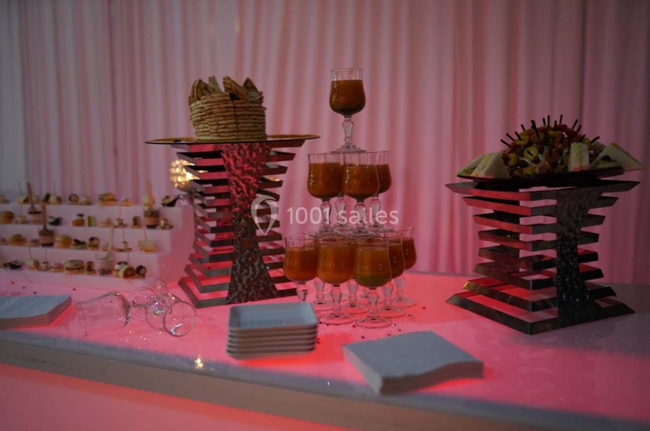 Buffet avec verrines de soupe, brochettes de fruits, biscuits et serviettes sur une table éclairée en rose.