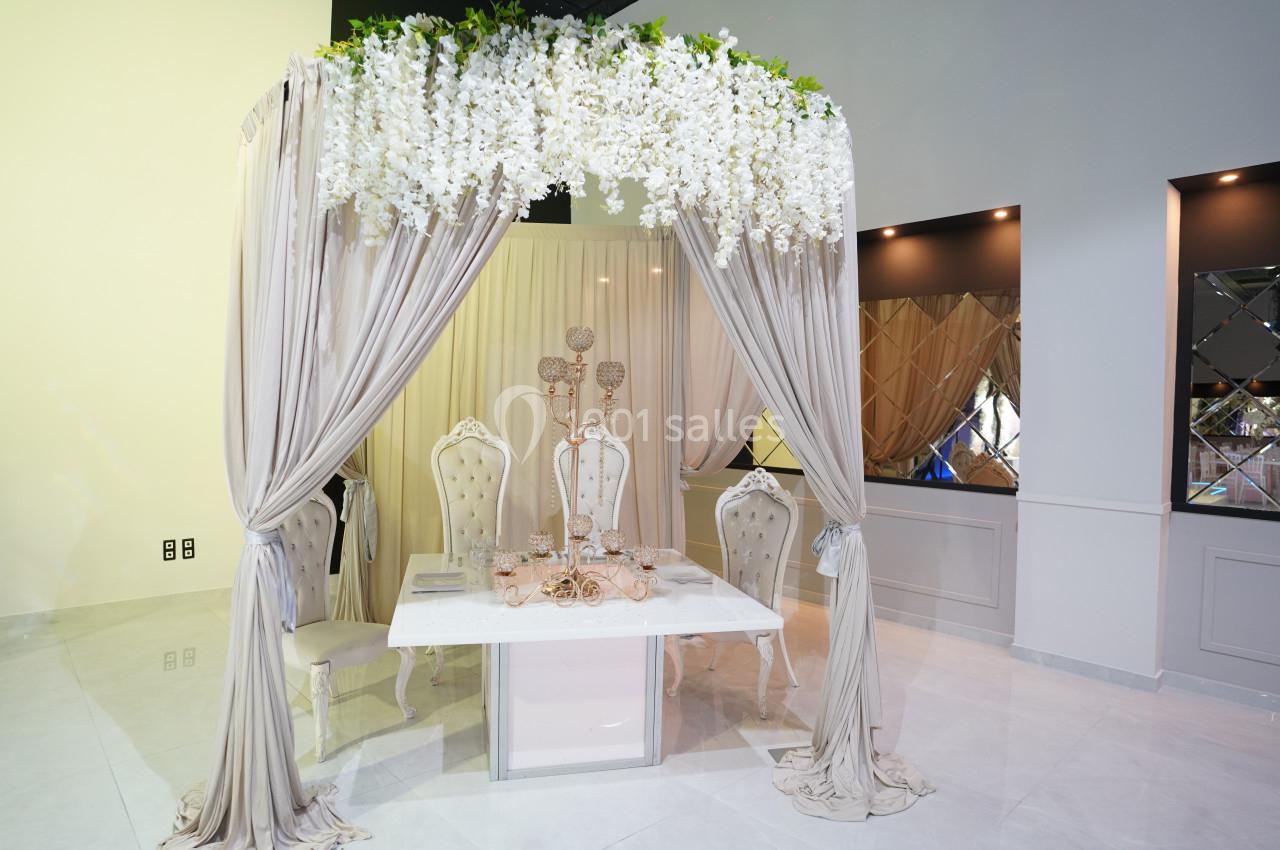 Table décorée avec des chaises élégantes sous une arche ornée de fleurs blanches et de rideaux beiges.