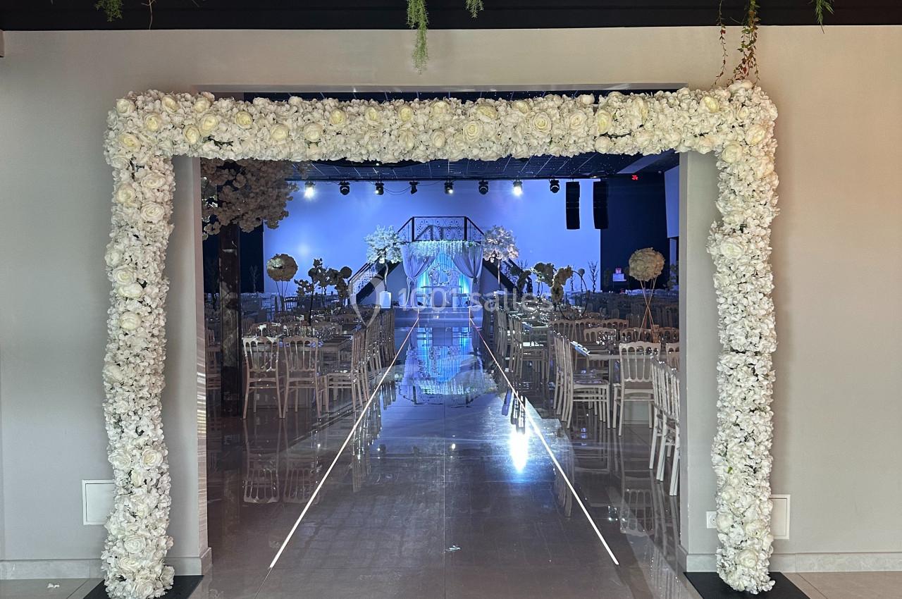 Allée centrale décorée de fleurs blanches menant à une scène illuminée, entourée de chaises transparentes et de verdure…