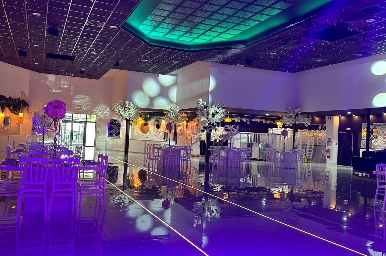 Salle de réception décorée avec des tables hautes, des chaises blanches et un éclairage violet et vert.