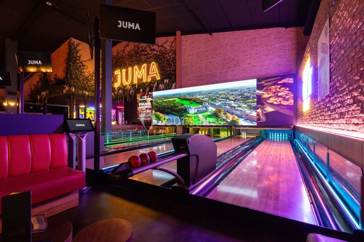 Piste de bowling moderne avec éclairage coloré, écran géant en fond et banquettes rouges sur le côté.