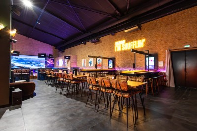 Salle de restaurant moderne avec bar illuminé, tables en bois, chaises colorées et décoration murale végétale.