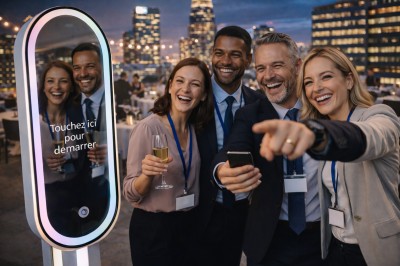 Un couple souriant en tenue de mariage pose devant une borne photo interactive lors d'une réception en extérieur.