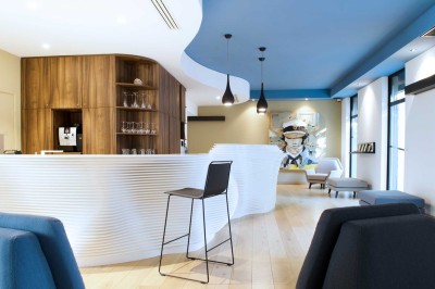 Chambre lumineuse avec lit double, table en bois, chaise beige, miroir et grande fenêtre avec stores.